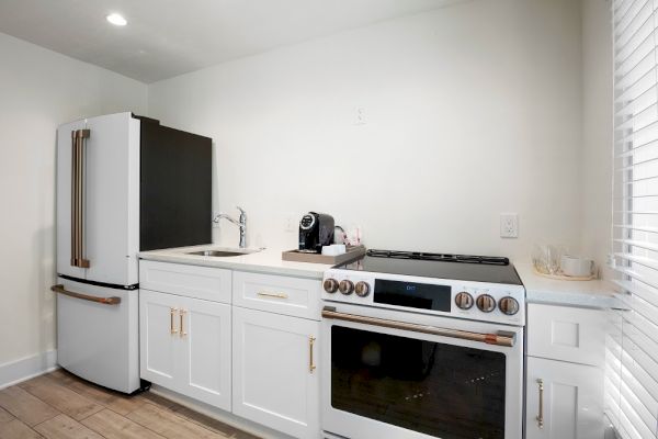 A modern kitchen with a refrigerator, sink, stove, and a coffee maker on the counter. White cabinets and countertops complement the look.