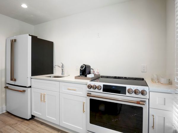 A modern kitchen with a refrigerator, sink, stove, and a coffee maker on the counter. White cabinets and countertops complement the look.