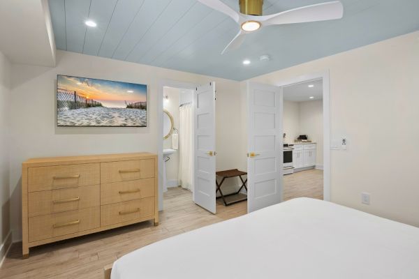 The image shows a bedroom with a dresser, artwork of a beach scene, a fan, and open doors leading to a bathroom and kitchen.