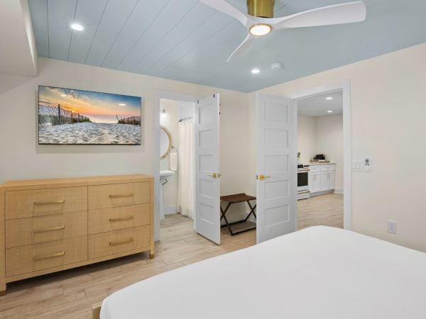 The image shows a bedroom with a dresser, artwork of a beach scene, a fan, and open doors leading to a bathroom and kitchen.