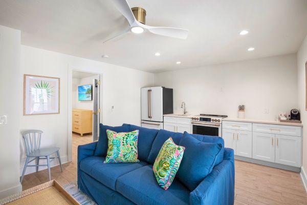 A cozy, modern living area with a blue sofa, colorful pillows, and a small kitchen, featuring white cabinets and light wood flooring.