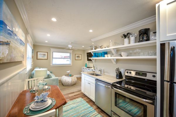 A cozy kitchen features open shelving, a small dining area, and modern appliances. It leads to a sitting area with a window.