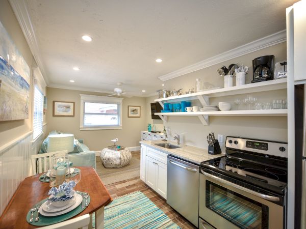 A cozy kitchen features open shelving, a small dining area, and modern appliances. It leads to a sitting area with a window.