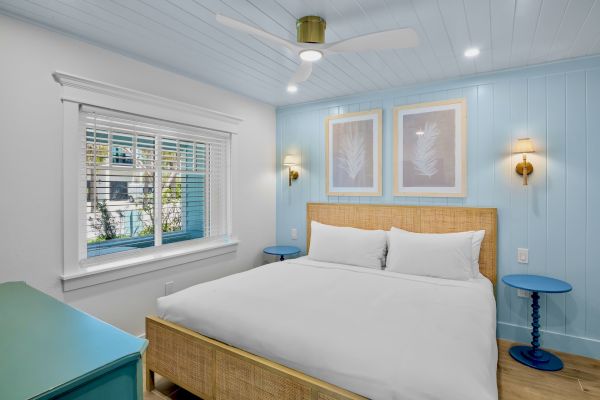 A cozy bedroom with light blue walls, a large bed, simple framed art, two bedside tables, and a ceiling fan above.