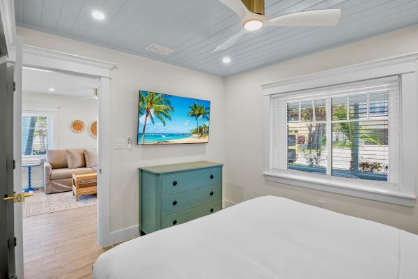A room with a bed, blue dresser, wall-mounted TV displaying a beach scene, a window, ceiling fan, and an open door to another room.