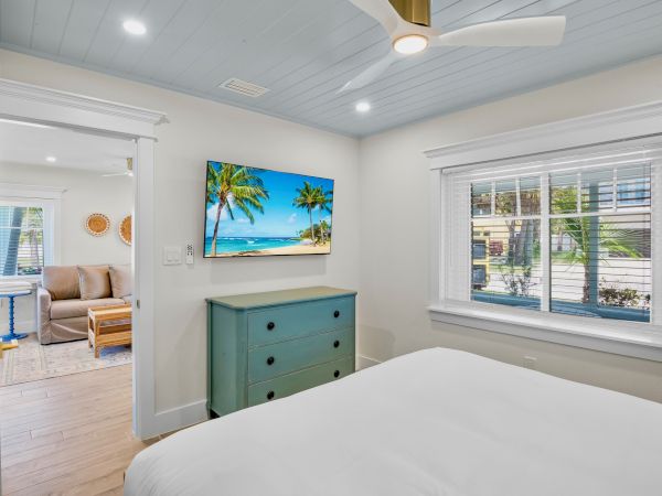 A room with a bed, blue dresser, wall-mounted TV displaying a beach scene, a window, ceiling fan, and an open door to another room.