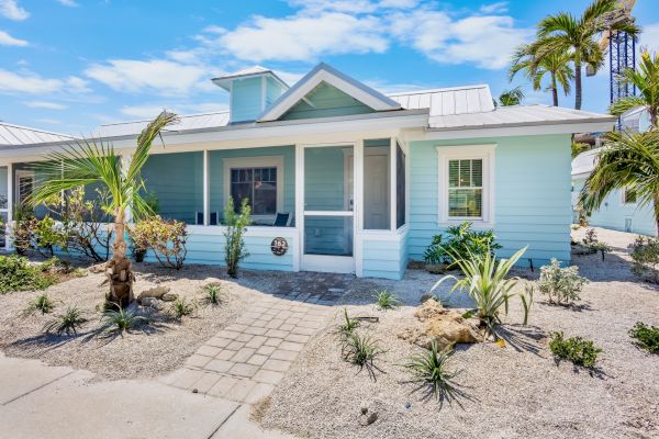 A light blue, beach-style house with a metal roof, screened porch, and sandy landscaping with palm trees and shrubs.
