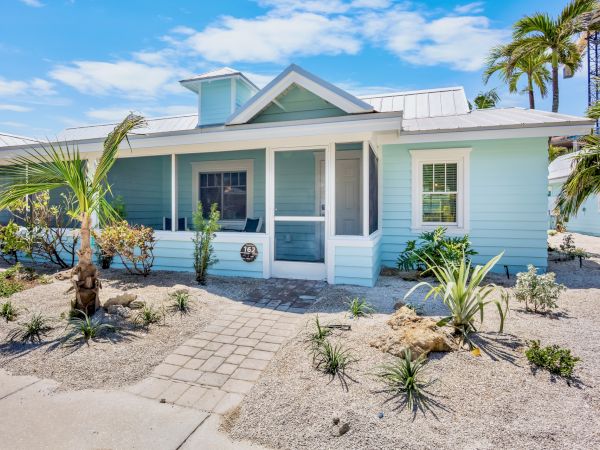 A light blue, beach-style house with a metal roof, screened porch, and sandy landscaping with palm trees and shrubs.