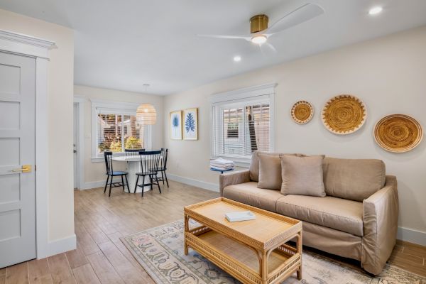 A cozy living room with a tan sofa, wicker coffee table, and wall decor; dining area with a round table and two chairs by the window.