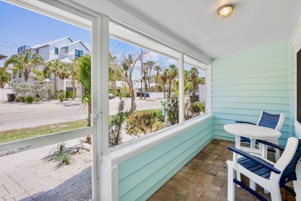 A porch with blue walls and white trim features a small table and two chairs, overlooking a street lined with palm trees and houses.