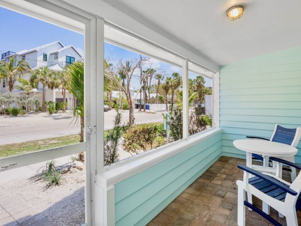 A porch with blue walls and white trim features a small table and two chairs, overlooking a street lined with palm trees and houses.