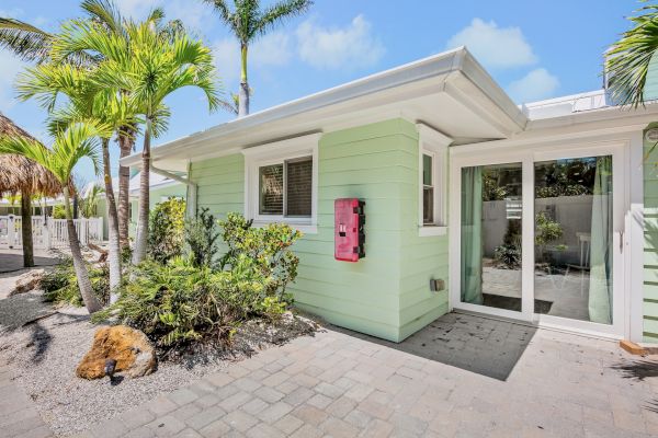 A small mint green house with palm trees, a pink mailbox, and sliding glass doors. The setting is sunny with a paver pathway.