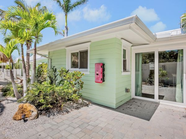 A small mint green house with palm trees, a pink mailbox, and sliding glass doors. The setting is sunny with a paver pathway.