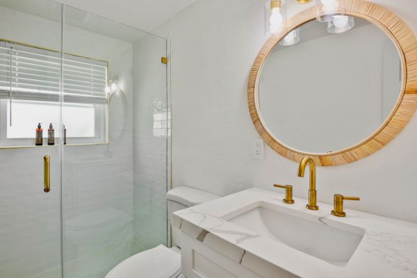 A modern bathroom with a circular mirror, gold fixtures, marble countertop, glass shower, and window with blinds.