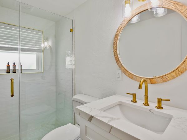 A modern bathroom with a circular mirror, gold fixtures, marble countertop, glass shower, and window with blinds.