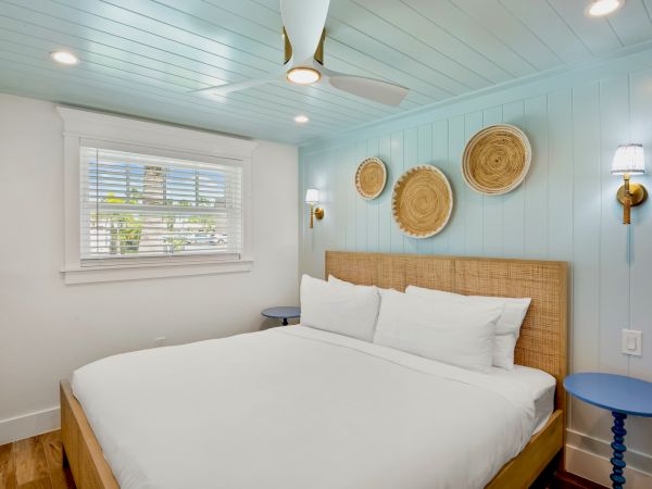 A cozy bedroom with a woven headboard, circular wall decor, blue side tables, and soft lighting, featuring a large window and ceiling fan.