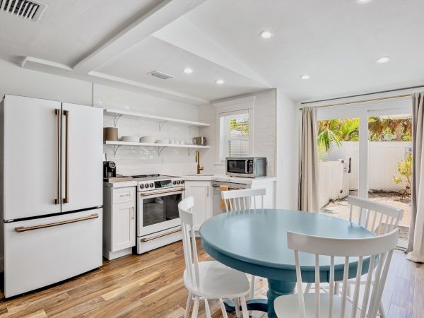 A modern kitchen with a white fridge, stove, open shelves, blue table, chairs, microwave, and a view of an outdoor area.