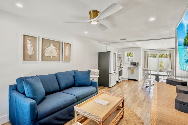 A modern living area with a blue sofa, kitchen, dining table, and tropical-themed decor and wall art, under a ceiling fan.