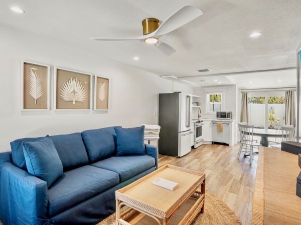 A modern living area with a blue sofa, kitchen, dining table, and tropical-themed decor and wall art, under a ceiling fan.