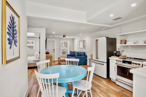 A cozy open-plan living area with a blue dining table, white chairs, kitchen appliances, a blue sofa, and artwork on the wall.