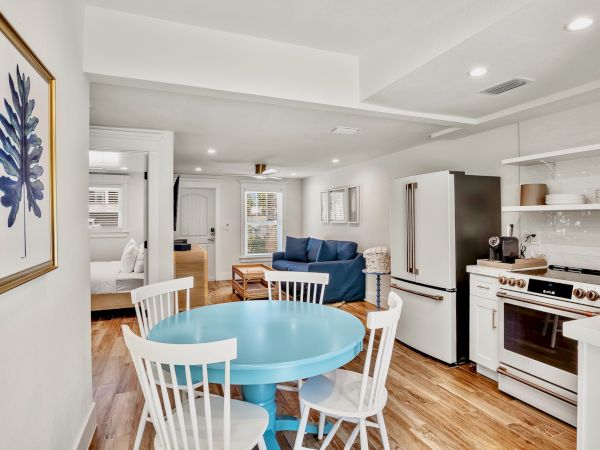 A cozy open-plan living area with a blue dining table, white chairs, kitchen appliances, a blue sofa, and artwork on the wall.