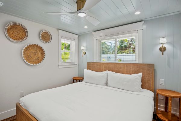A cozy bedroom features a large bed with white linens, woven wall decor, bedside tables, and bright windows for a serene atmosphere.