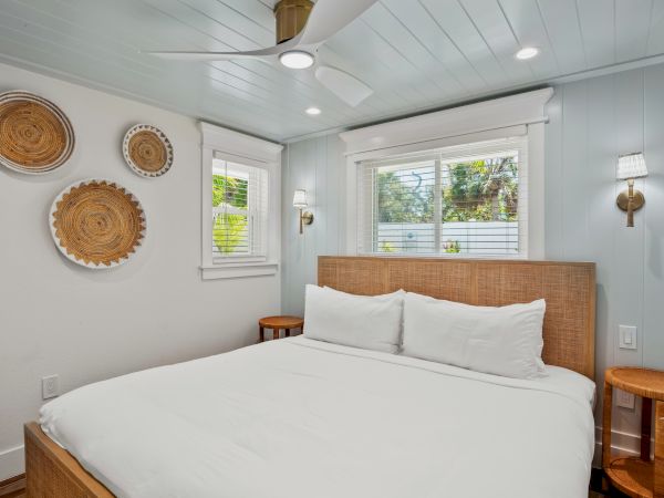 A cozy bedroom features a large bed with white linens, woven wall decor, bedside tables, and bright windows for a serene atmosphere.