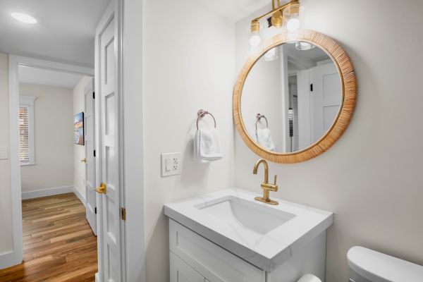 A modern bathroom with a round mirror, gold faucet, white vanity, and towel ring. Wood floors extend into the hallway.