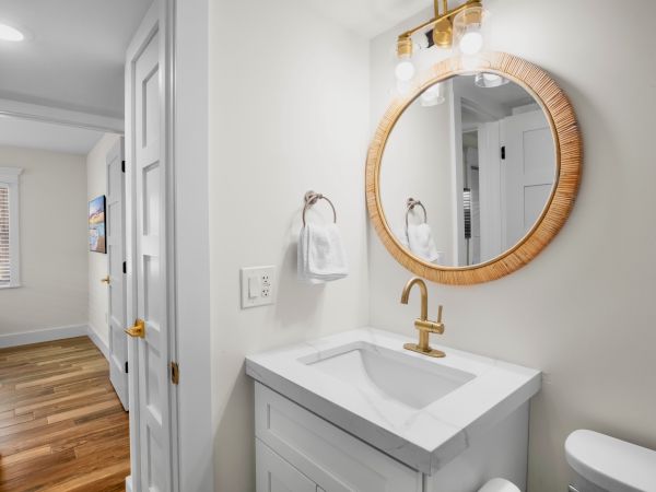 A modern bathroom with a round mirror, gold faucet, white vanity, and towel ring. Wood floors extend into the hallway.