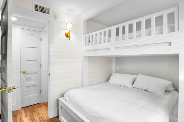 A room with white bunk beds, a wooden floor, a closed door, and a wall-mounted light fixture.