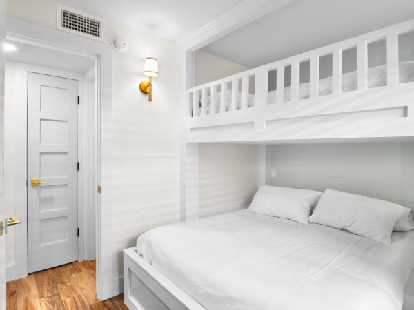A room with white bunk beds, a wooden floor, a closed door, and a wall-mounted light fixture.