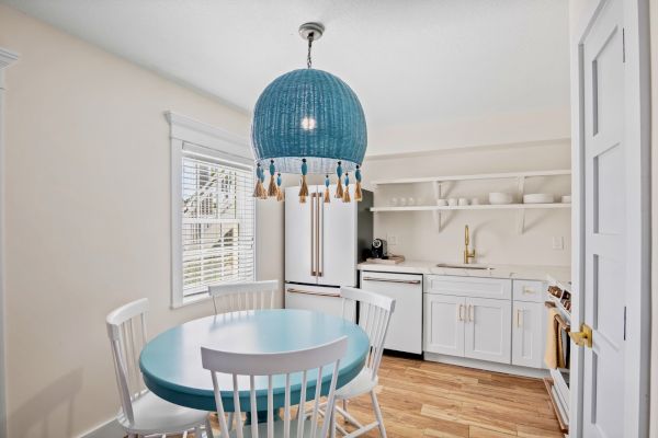 A bright kitchen with a blue table, white chairs, and a blue tassel chandelier. White cabinets, a sink, and open shelves are visible.
