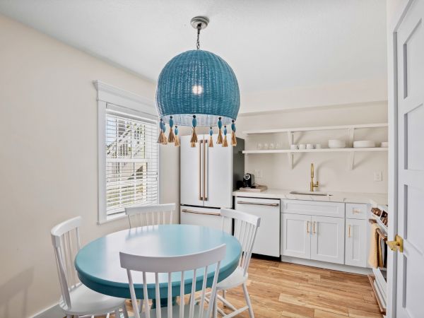 A bright kitchen with a blue table, white chairs, and a blue tassel chandelier. White cabinets, a sink, and open shelves are visible.