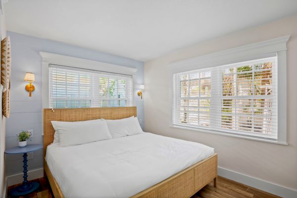 A bright bedroom with a double bed, wooden frame, two windows with blinds, wall lamps, and a small table with a plant.