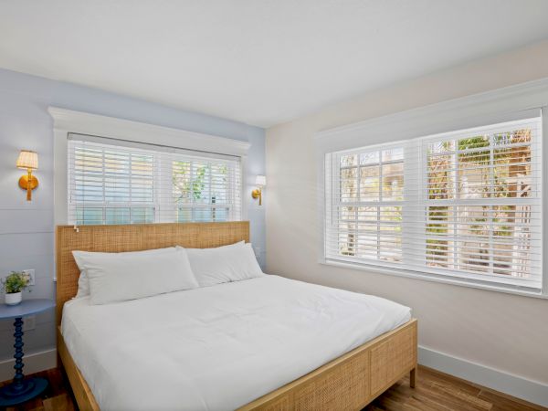 A bright bedroom with a double bed, wooden frame, two windows with blinds, wall lamps, and a small table with a plant.