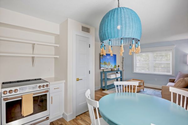 A cozy room with a blue round table, white chairs, teal lamp, TV, and sofa; kitchen area features a stove and minimal shelving.