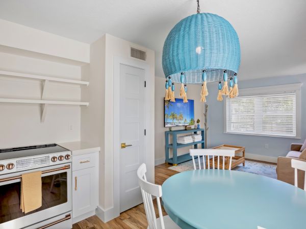A cozy room with a blue round table, white chairs, teal lamp, TV, and sofa; kitchen area features a stove and minimal shelving.