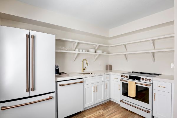 Modern kitchen with white cabinets, gold handles, open shelves, a fridge, oven, sink, and wooden floor. Clean and minimal design.