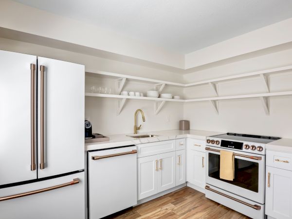 Modern kitchen with white cabinets, gold handles, open shelves, a fridge, oven, sink, and wooden floor. Clean and minimal design.