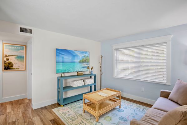 A cozy room features a sofa, TV, wicker coffee table, decorative rug, and window with blinds. A wall poster adds a beachy vibe.