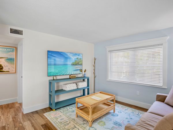 A cozy room features a sofa, TV, wicker coffee table, decorative rug, and window with blinds. A wall poster adds a beachy vibe.
