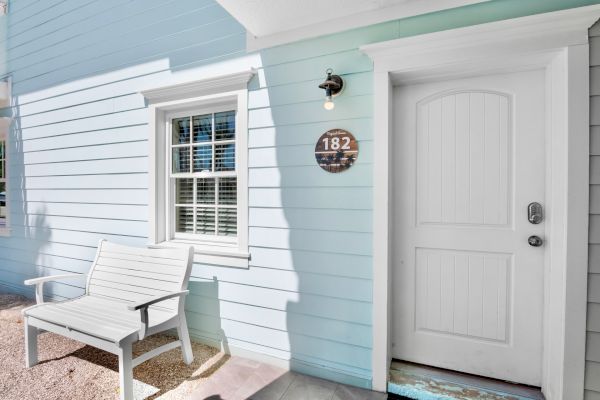 A light blue house exterior with a white door, a window with blinds, a wall-mounted light, and a wooden bench. The number 182 is displayed.