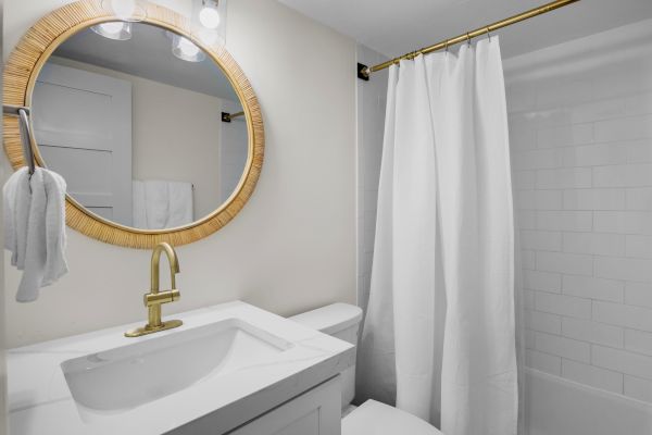 A bathroom with a round mirror, white towels, a sink, gold faucet, and a shower with a white curtain ends the scene.