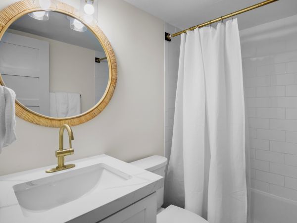 A bathroom with a round mirror, white towels, a sink, gold faucet, and a shower with a white curtain ends the scene.