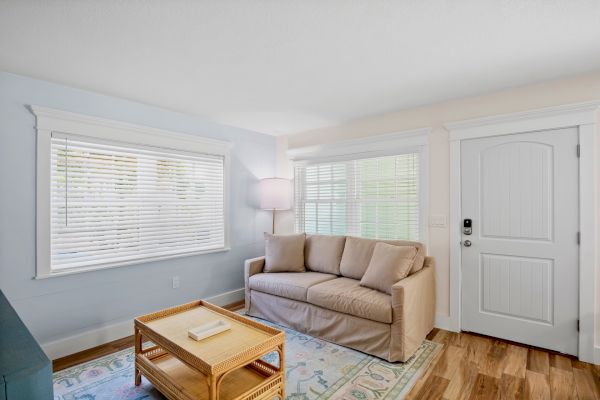 A cozy living room with a beige sofa, wicker coffee table, standing lamp, large windows with blinds, and a white door on wooden flooring.