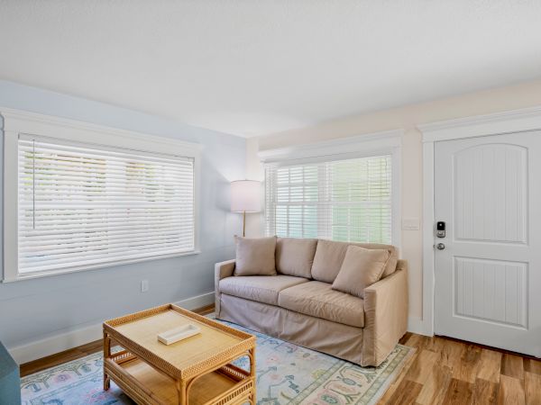 A cozy living room with a beige sofa, wicker coffee table, standing lamp, large windows with blinds, and a white door on wooden flooring.