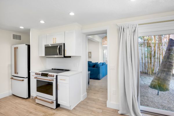 A modern kitchen with white cabinets, stainless steel appliances, and wood floors; a living area and outdoor view are visible.