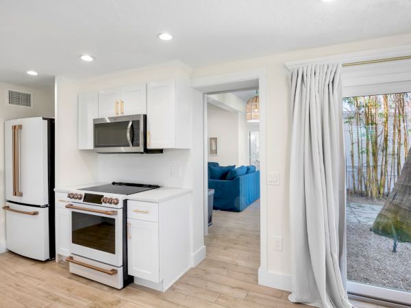 A modern kitchen with white cabinets, stainless steel appliances, and wood floors; a living area and outdoor view are visible.