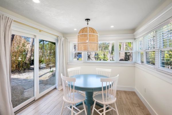 A bright dining nook with a round blue table, four white chairs, wicker pendant light, large windows, and sliding glass doors to a garden.