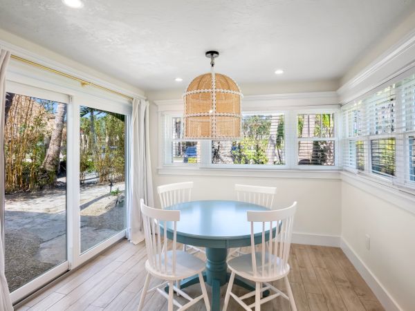 A bright dining nook with a round blue table, four white chairs, wicker pendant light, large windows, and sliding glass doors to a garden.